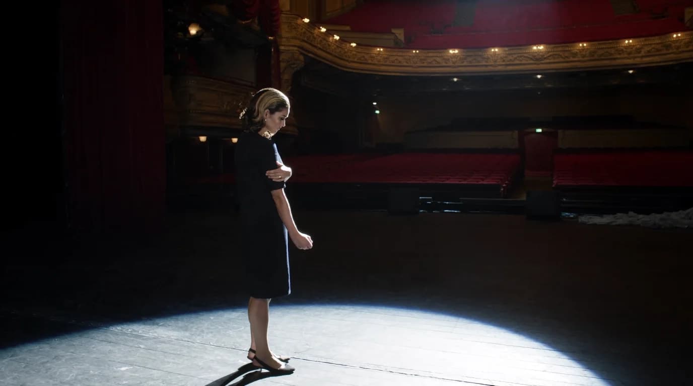 A woman stands alone in a spotlight on a theatre stage, looking down, with rows of empty red seats behind her